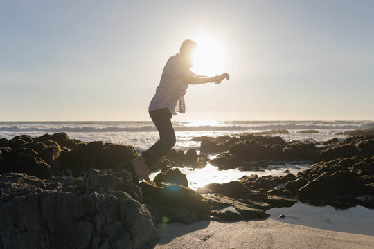 Man jumping barefoot between jagged rocks on beach at low tide wearing striped shirt and trousers - Powered by Adobe