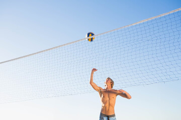 Athletic male wearing swim shorts spiking volleyball over net on beach under blue sky, copy space