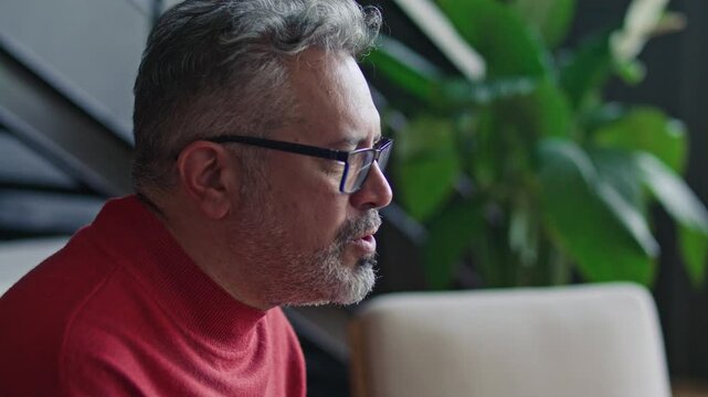 Middle-aged man in glasses talking openly during therapy session in modern office with green plants in background. Profile close-up view