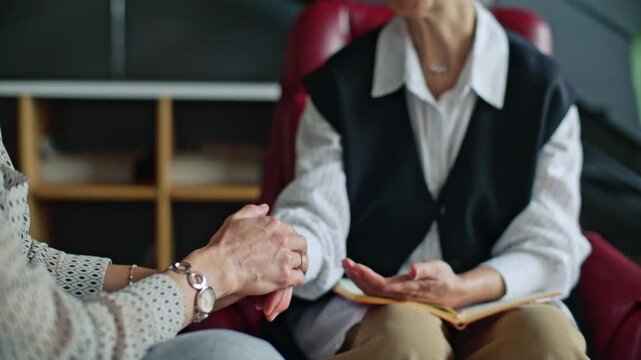 Empathic senior therapist gently holding hands of female patient while and talking calmly to reassure her during psychology consultation in office. Tilt-down shot