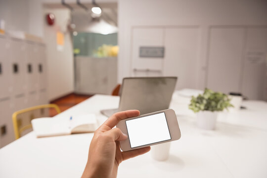 Hand holding smartphone over office desk, featuring blank screen with notebook and laptop