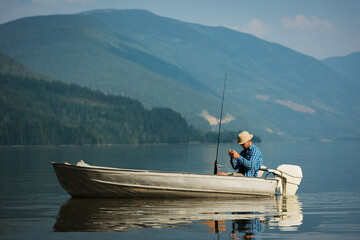 Male angler wearing bucket hat baiting fishing line on aluminum boat at mountain lake, copy space