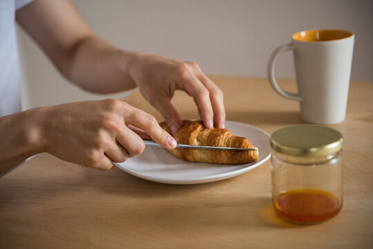 Asian man slicing golden-brown croissant on ceramic plate at dining table with jam jar, cream mug