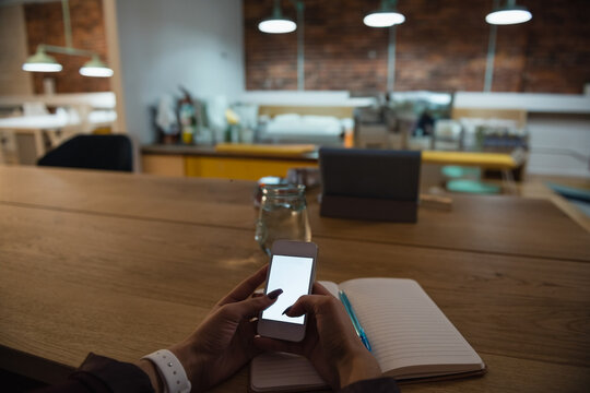 Hands holding smartphone above open notebook with pen on table in cafe with jar and tablet