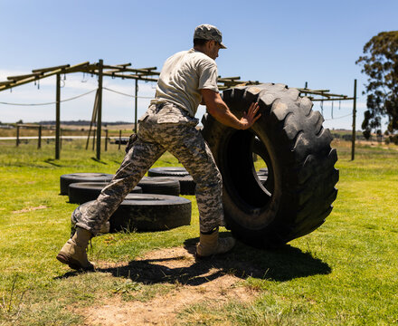 Male soldier pushing large tractor tire near obstacle beams with tires on military training ground - Powered by Adobe