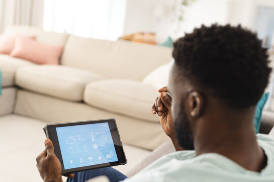 African American man sitting in living room holding tablet showing smart home app, copy space