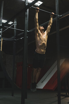 Shirtless man gripping pull-up bar on metal rig in gym under fluorescent lights with striped wall