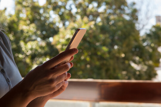 Woman holding smartphone and scrolling screen on wood balcony railing overlooking trees, copy space
