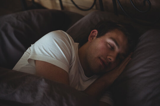 Man in his twenties lying on bed in bedroom wearing t-shirt under charcoal duvet by headboard