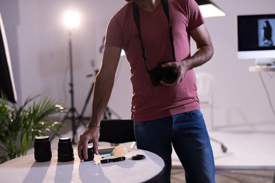 Male photographer wearing red t-shirt and jeans arranging DSLR camera and lenses on table in studio