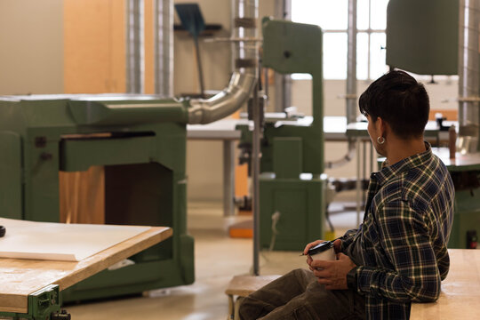 Hispanic man in woodshop sitting on workbench holding mug gazing toward green sander, copy space - Powered by Adobe