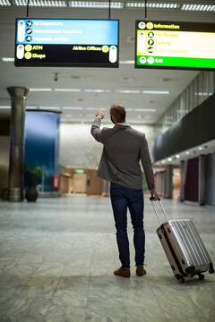 White middle-aged man pointing at overhead signs while pulling wheeled suitcase at airport hall