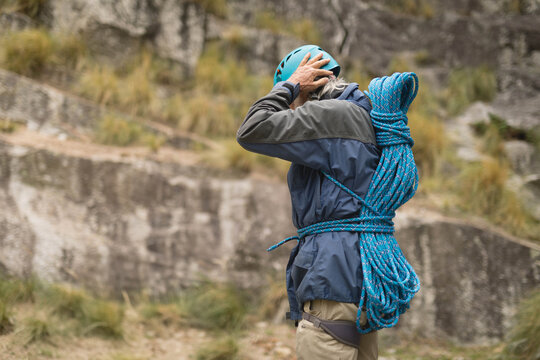 Senior man adjusting teal climbing helmet with bright blue rope harness on rocky hill, copy space