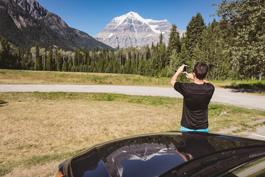 Adult male capturing snow-capped mountain on smartphone by parked car at grassy pullout, copy space