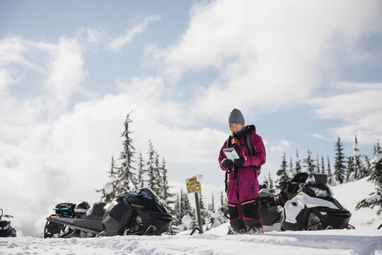 Woman examining map while standing on snow-covered slope between snowmobiles and carrying backpack