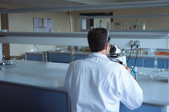 Mature adult male scientist in lab coat leaning toward microscope, examining bottles at lab bench