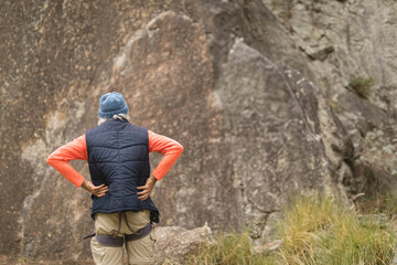 Senior man planning ascent wearing knit beanie, quilted vest, harness on grassy slope by rock face