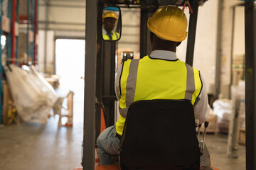 African American male driving forklift down warehouse aisle wearing safety vest, hardhat