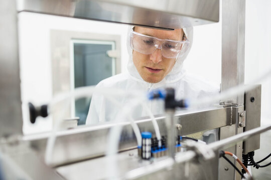 Male lab technician wearing coverall inspecting vials tubing and connectors on machine in lab