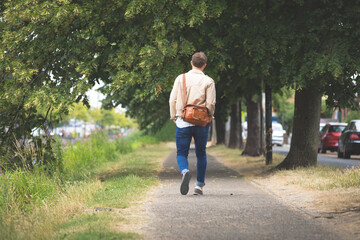 Mid adult male walking under green canopy on suburban sidewalk carrying brown leather messenger bag