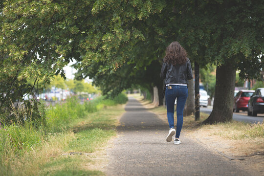 Mid adult woman walking on tree-lined paved path beside cars with crossbody bag and smartphone