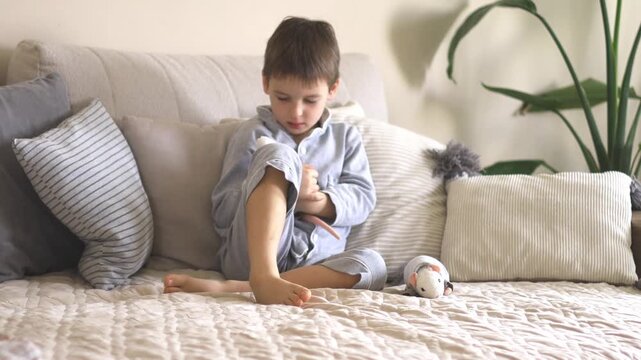 A little European boy of six years old in gray pajamas sits on a sofa and plays with soft toys