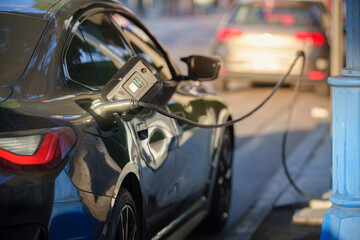 Electric cars charging on city street at sunset. Focus on EV charging station cable plugged into charging port. Shallow DOF.