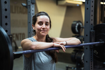 Latin woman smiling, leaning on a barbell during her strength workout in the gym, showing fitness...