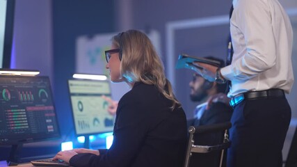 In a modern data analysis center, a female analyst reviews performance dashboards across multiple screens while a manager oversees, collaborating to derive insights from business data. - Powered by Adobe