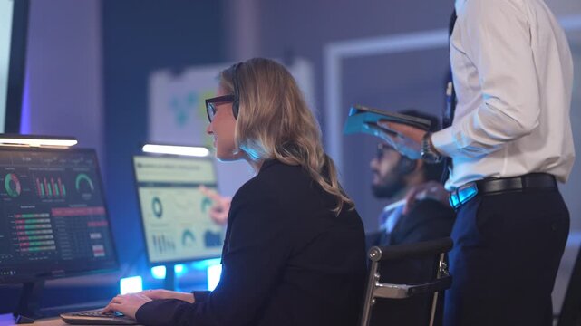 In a modern data analysis center, a female analyst reviews performance dashboards across multiple screens while a manager oversees, collaborating to derive insights from business data.