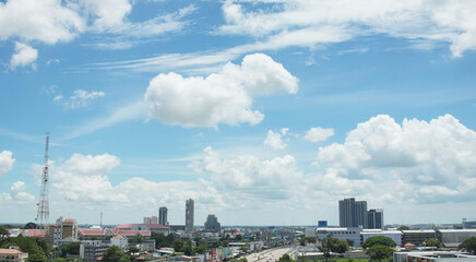 Panoramic view of cityscape with tall buildings communication towers and residential areas beneath vivid blue sky filled with fluffy white clouds.
