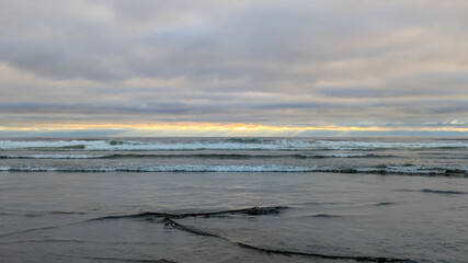 Side view from a wet beach with an emphasis on the small waves rolling in, out westward towards the pacific ocean, under a majestic and complicated cloud-scape near dusk.