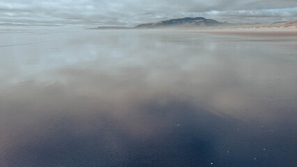 View northward along a beach towards a distant ridge covered in a dark pine forest, with the cloudy and  mysterious sky reflected on the wet sand. 