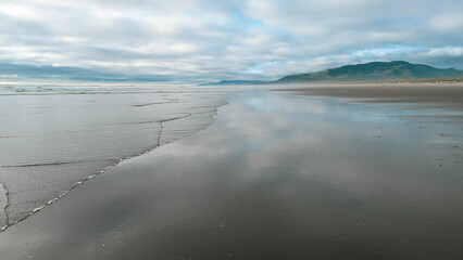 View northward along a beach towards a distant ridge covered in a dark pine forest, with the cloudy and  mysterious sky reflected on the wet sand.
