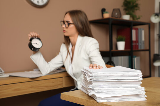 Young businesswoman with alarm clock and papers trying to meet deadline in office, closeup