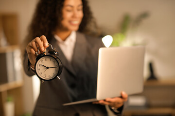 Young businesswoman with alarm clock and laptop meeting deadline in office at night, closeup
