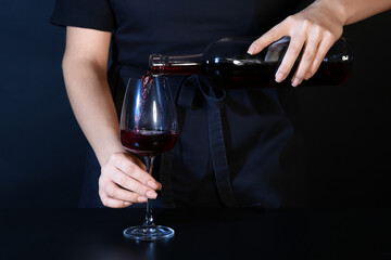 Female sommelier pouring red wine from jug into glass on black background