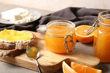 Wooden board with slices of bread and jar of sweet orange jam on grey background