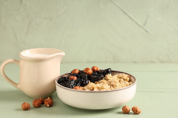 Bowl of tasty oatmeal with prunes, hazelnuts and jug of milk on green wooden background