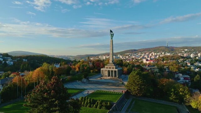 Drone view of Slavin War Memorial in Bratislava, Slovakia, surrounded by autumn foliage and panoramic cityscape under a clear blue sky.