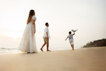 Happy family walking on the beach at sunset, child playing with toy airplane, symbolizing family bonding, travel, freedom, and carefree childhood moments.