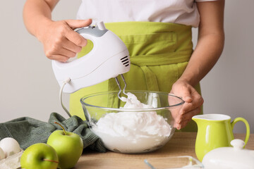 Woman whipping egg albumen with mixer at table on light background, closeup