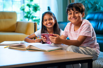 Indian Asian siblings kids focused on studying with books and smartphone together in modern home