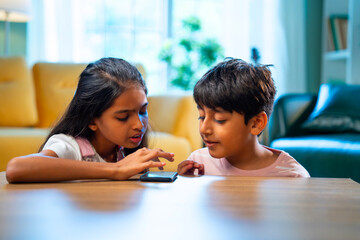 Indian Asian siblings kids using smartphones to play games while enjoying leisure time at home