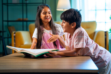 Obraz premium Indian Asian siblings kids reading book and studying together at home in a modern family lifestyle