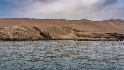 An island in the ocean. Sandy slopes without vegetation. Blue sky, clouds. The surface of the sea is in the foreground. Peru. Paracas. Ballestas Islands