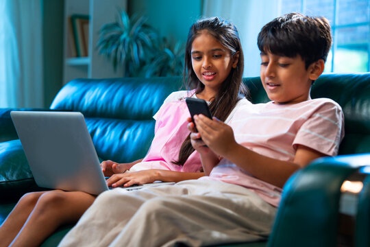 Indian kids sitting on sofa using laptop and smartphone, siblings enjoy digital time at home - Powered by Adobe