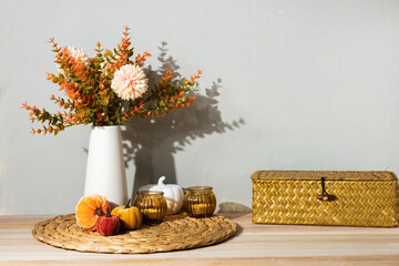 A white vase with autumn flowers stands on a woven placemat, decorated with amber glass candle holders and a white pumpkin. The cozy still life creates a warm seasonal home atmosphere.