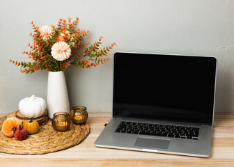 A laptop stands on a wooden desk next to a notebook, autumn flowers in a vase, candles, and decorative pumpkins. The cozy workspace creates a warm seasonal atmosphere for work or study.