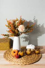 A white vase with autumn flowers stands on a woven placemat, decorated with amber glass candle holders and a white pumpkin. The cozy still life creates a warm seasonal home atmosphere.